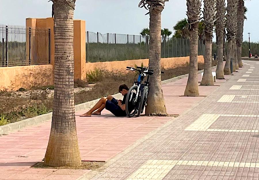 Joven sentado en el paseo marítimo junto a su bicicleta y palmeras