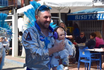 Hombre con disfraz brillante sostiene a un bebé en un desfile de carnaval