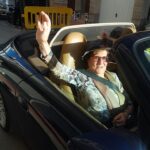 Mujer mayor saludando desde un coche descapotable en la calle.
