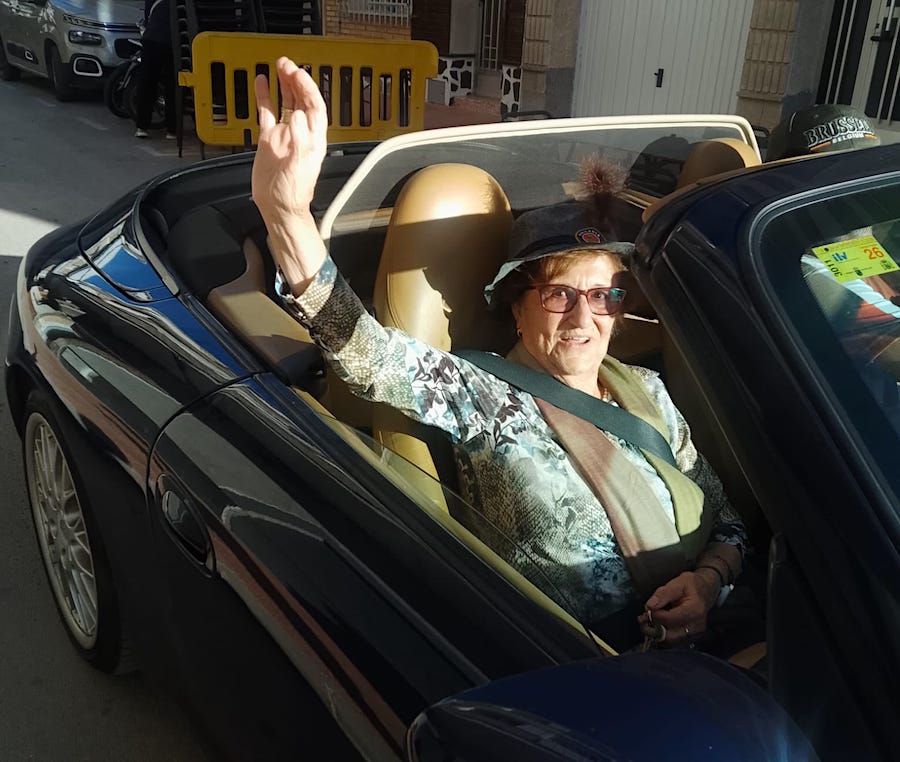 Mujer mayor saludando desde un coche descapotable en la calle.